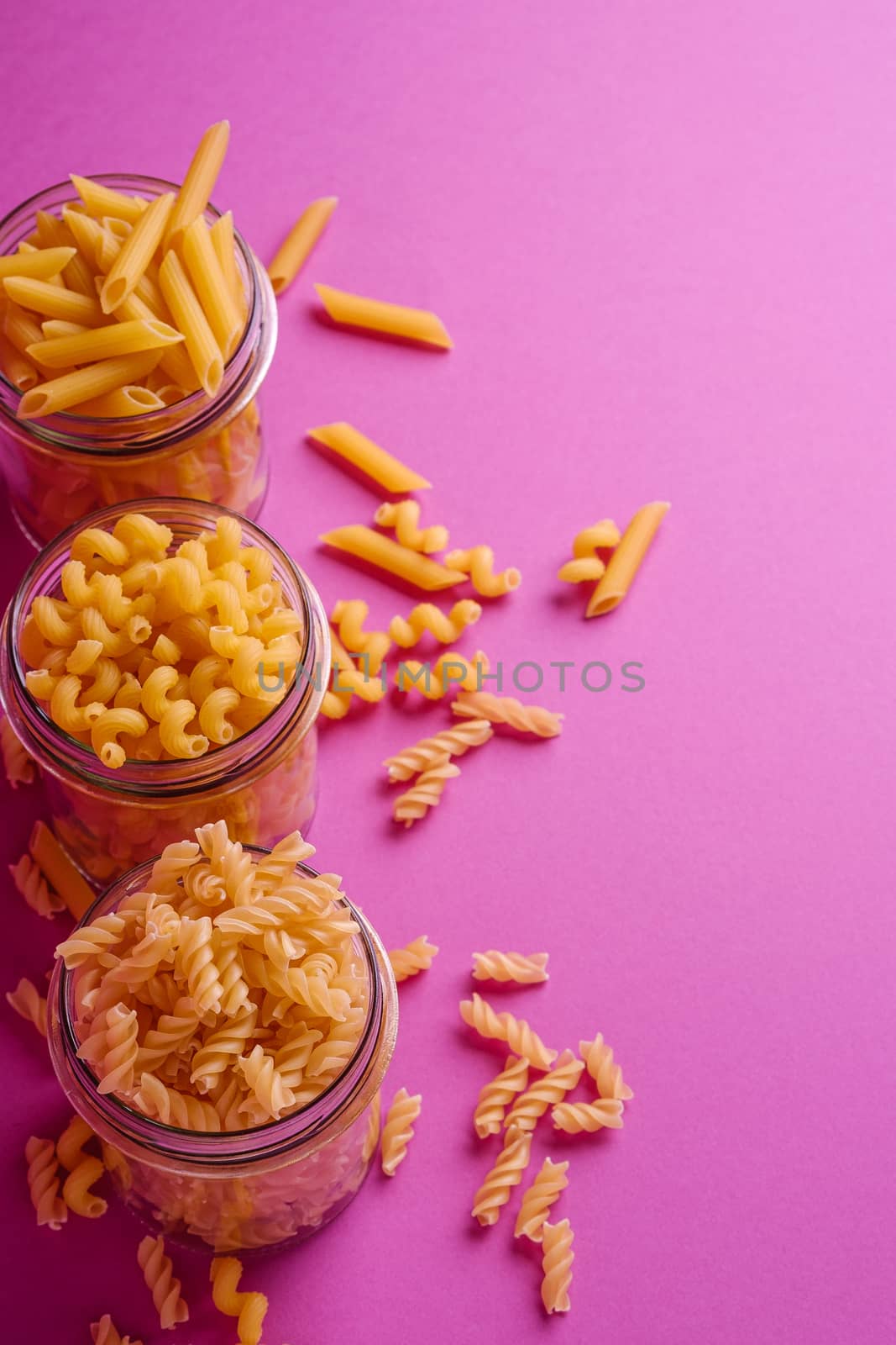 Three glass jars with variety of uncooked golden wheat pasta on minimal pink background, angle view