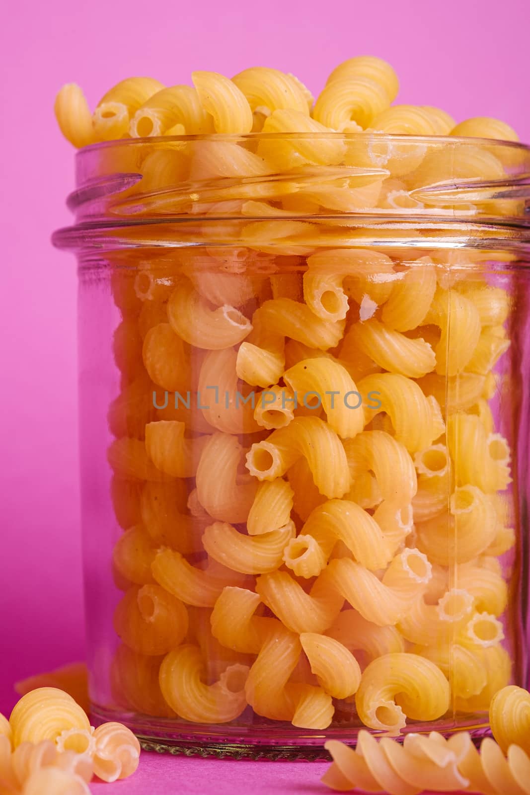 One glass jar with cavatappi uncooked golden wheat curly pasta on minimal pink background, angle view macro