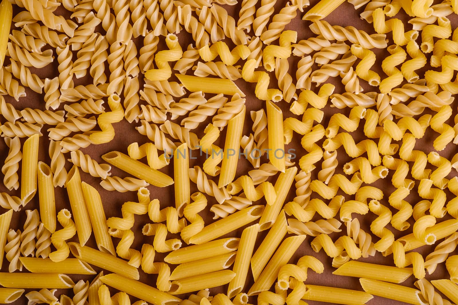Scattered variety of uncooked golden wheat pasta on minimal brown background, top view