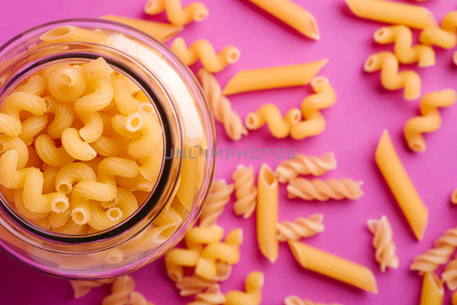 One glass jar with variety of uncooked golden wheat pasta on minimal pink background, top view macro
