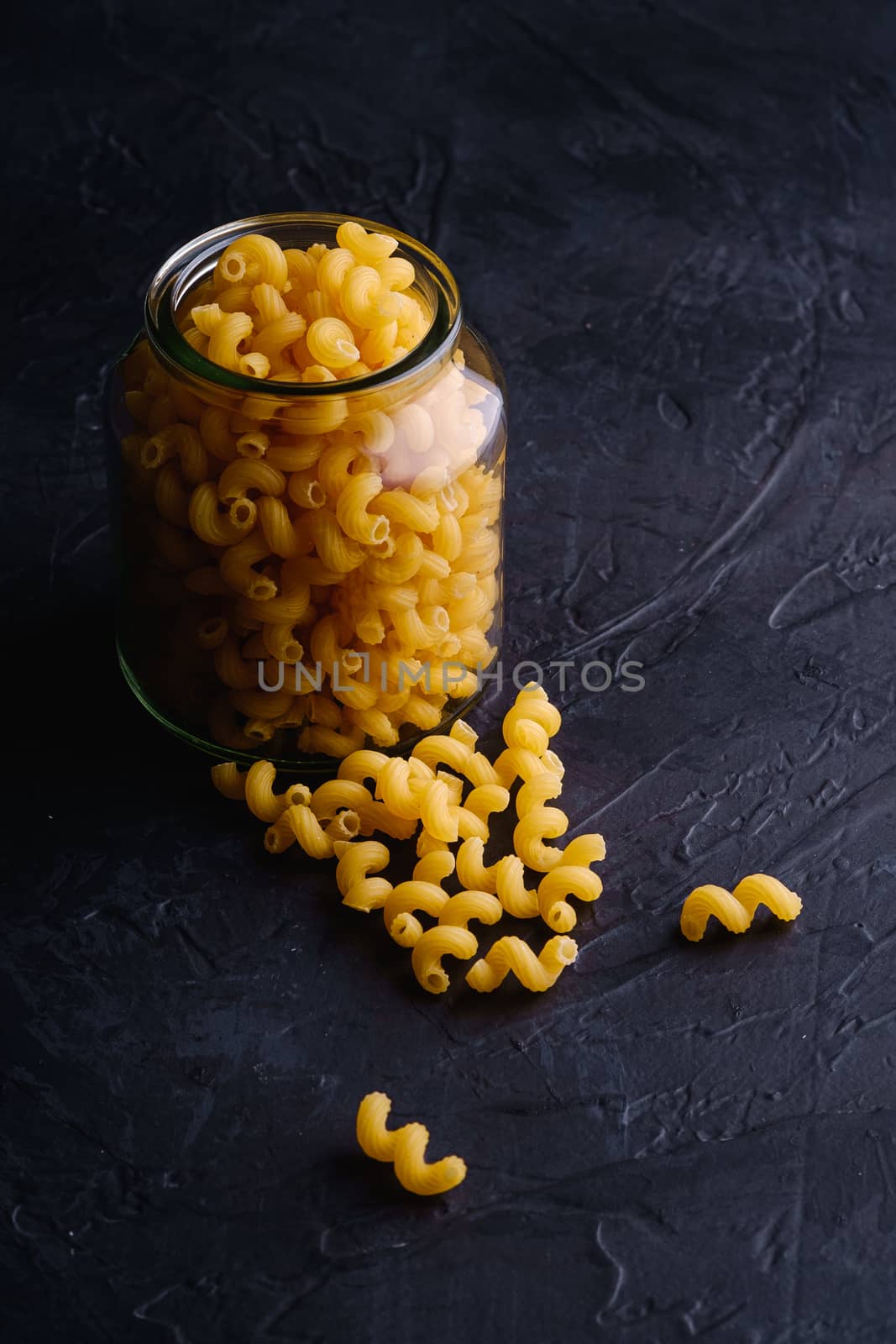 One glass jar with cavatappi uncooked golden wheat curly pasta near to scattered macaroni on textured dark black background, angle view