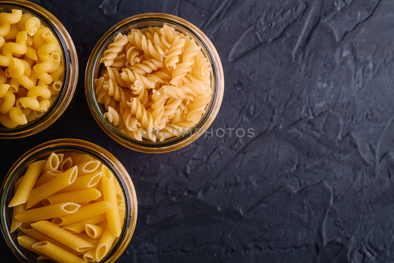 Three glass jars with variety of uncooked golden wheat pasta on dark black textured background, top view copy space