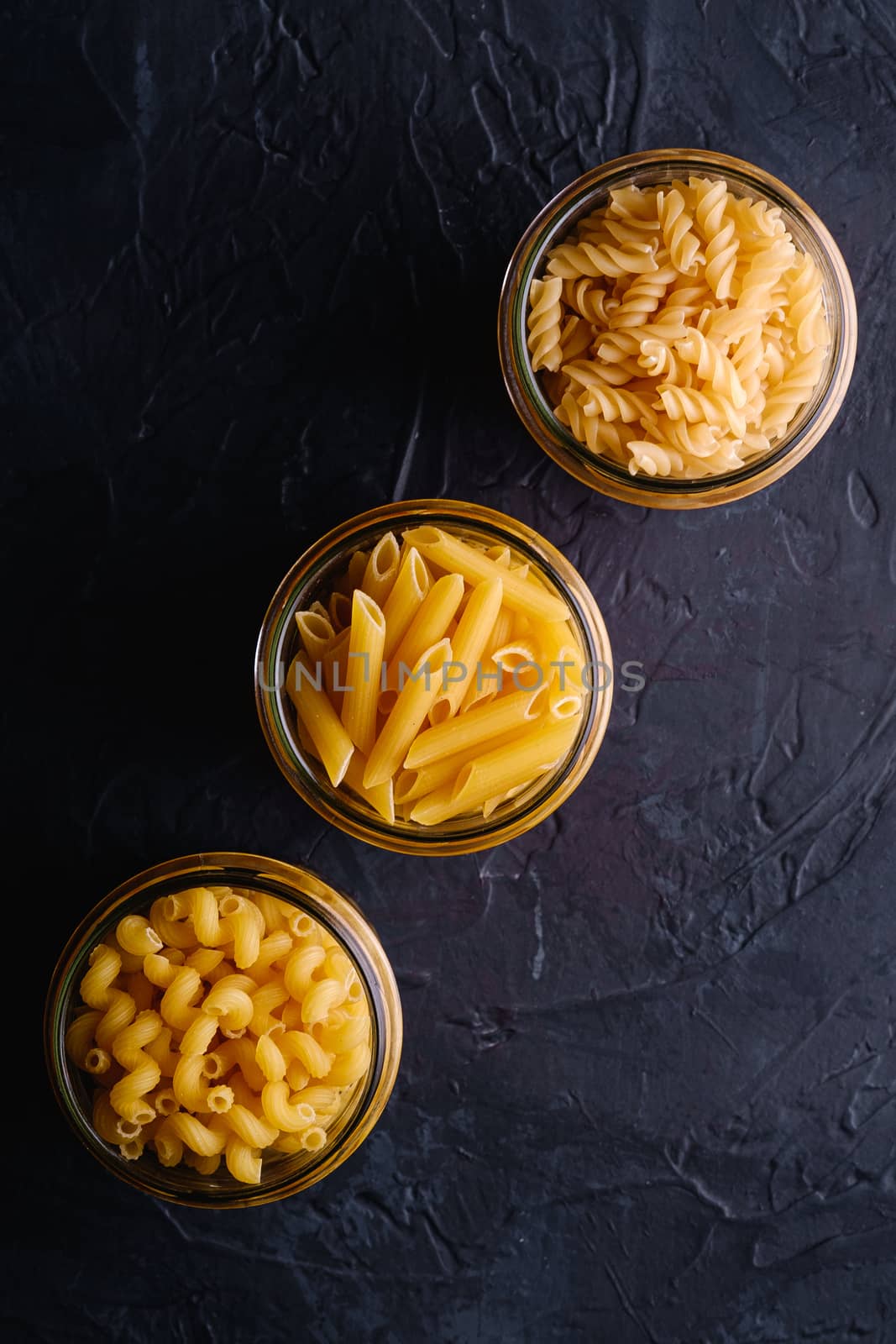 Three glass jars in row with variety of uncooked golden wheat pasta on dark black textured background, top view