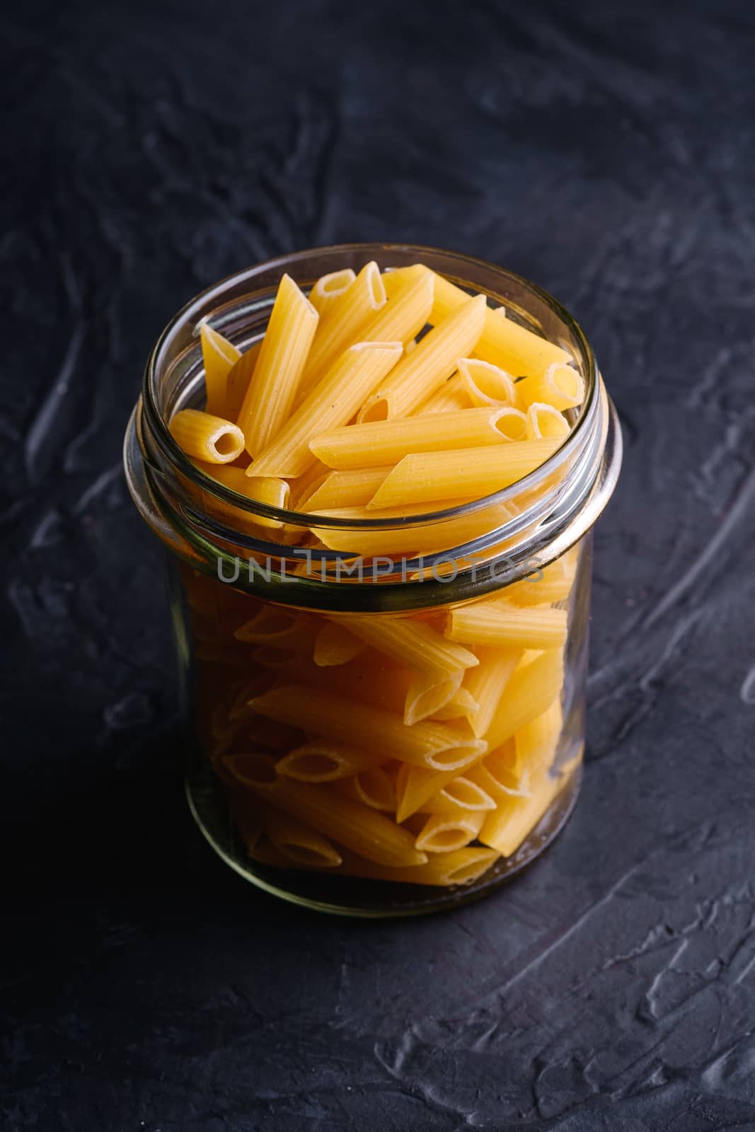 One glass jar with penne uncooked golden wheat tube pasta on textured dark black background, angle view