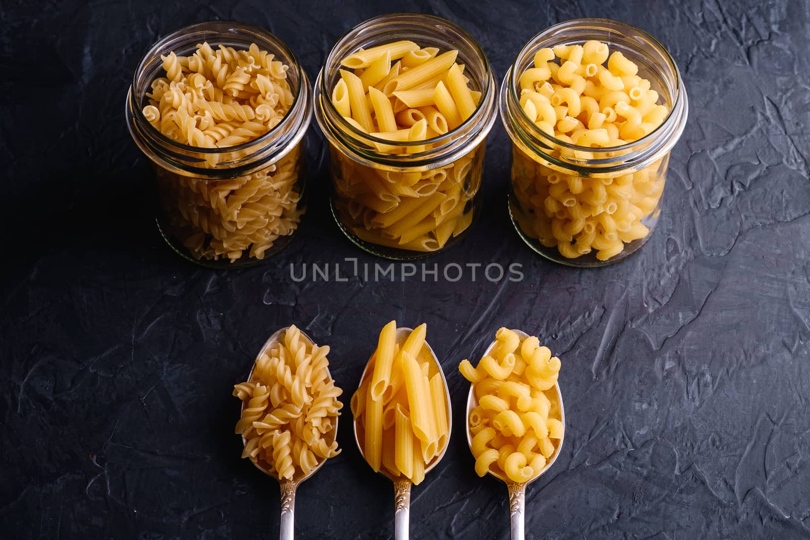 Three cutlery spoons and glass jars with variety of uncooked golden wheat pasta on dark black textured background, angle view