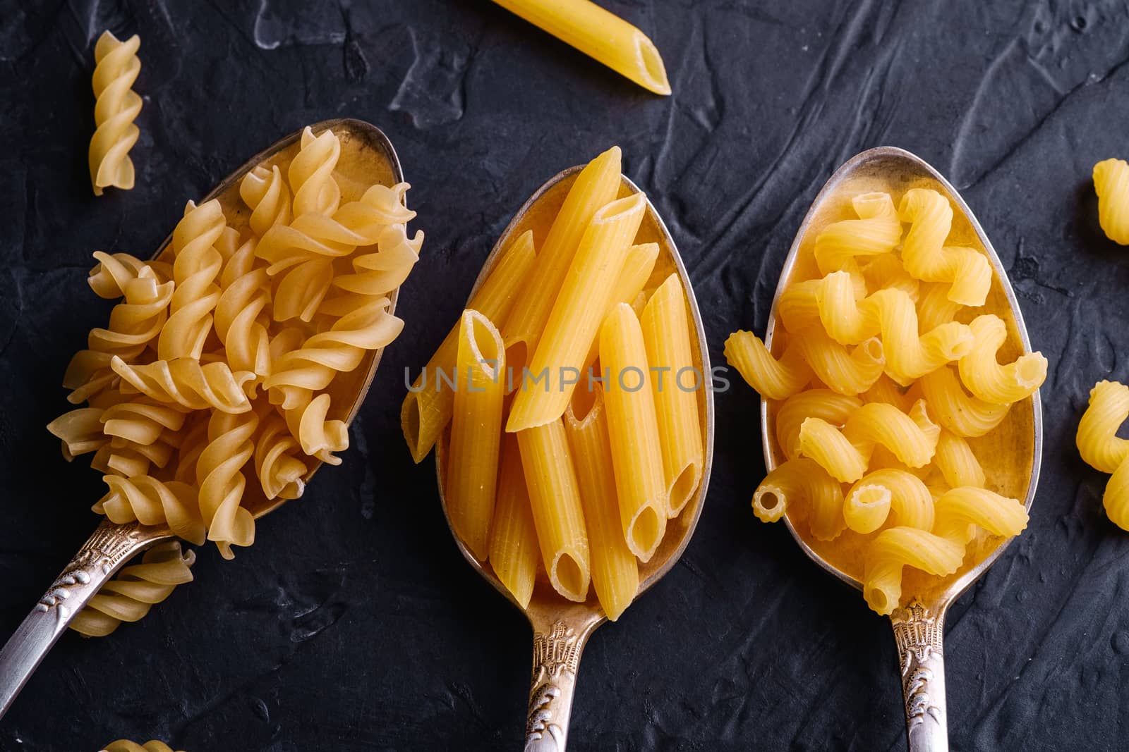 Three cutlery spoons with variety of uncooked golden wheat pasta on dark black textured background, top view macro