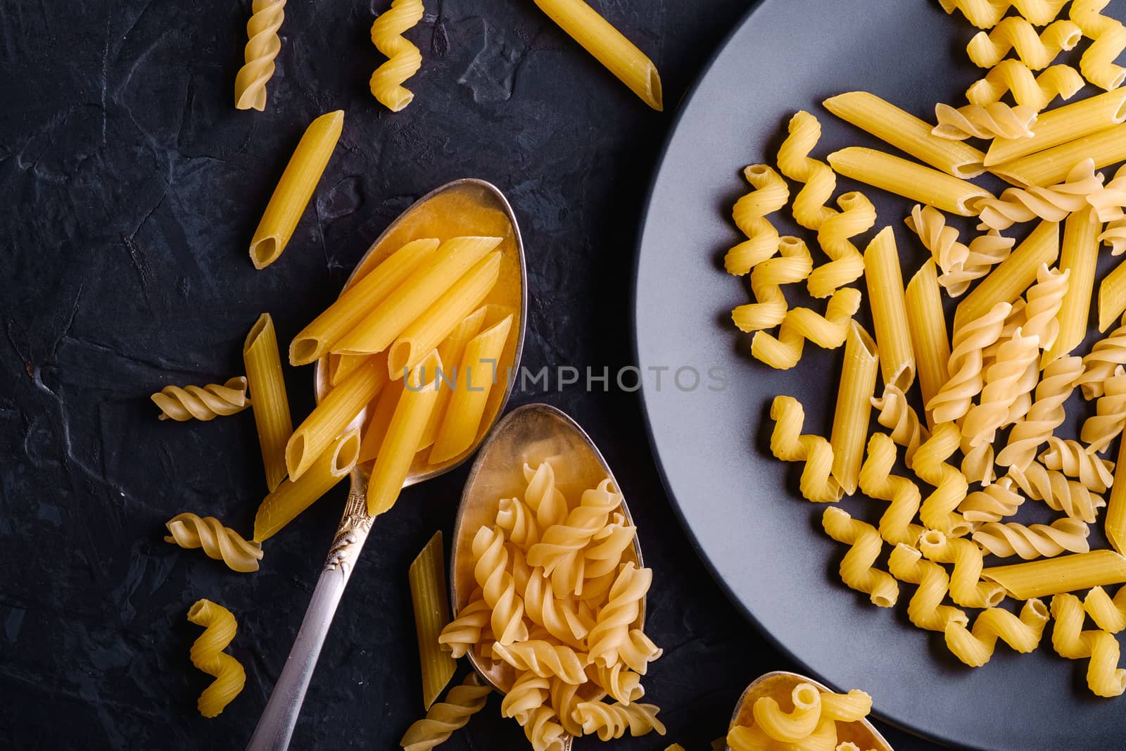 Three cutlery spoons and plate with variety of uncooked golden wheat pasta on dark black textured background, top view macro