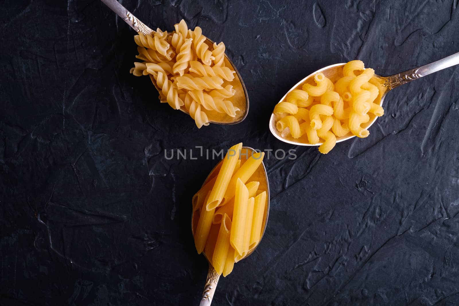 Three cutlery spoons with variety of uncooked golden wheat pasta on dark black textured background, top view