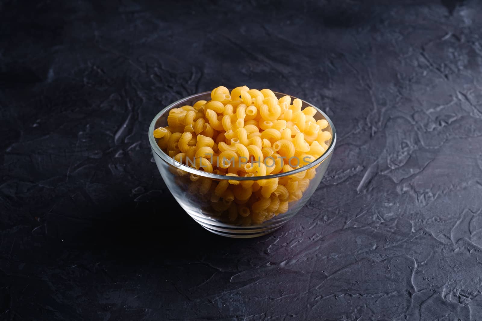 Glass bowl with cavatappi uncooked golden wheat curly pasta on textured dark black background, angle view