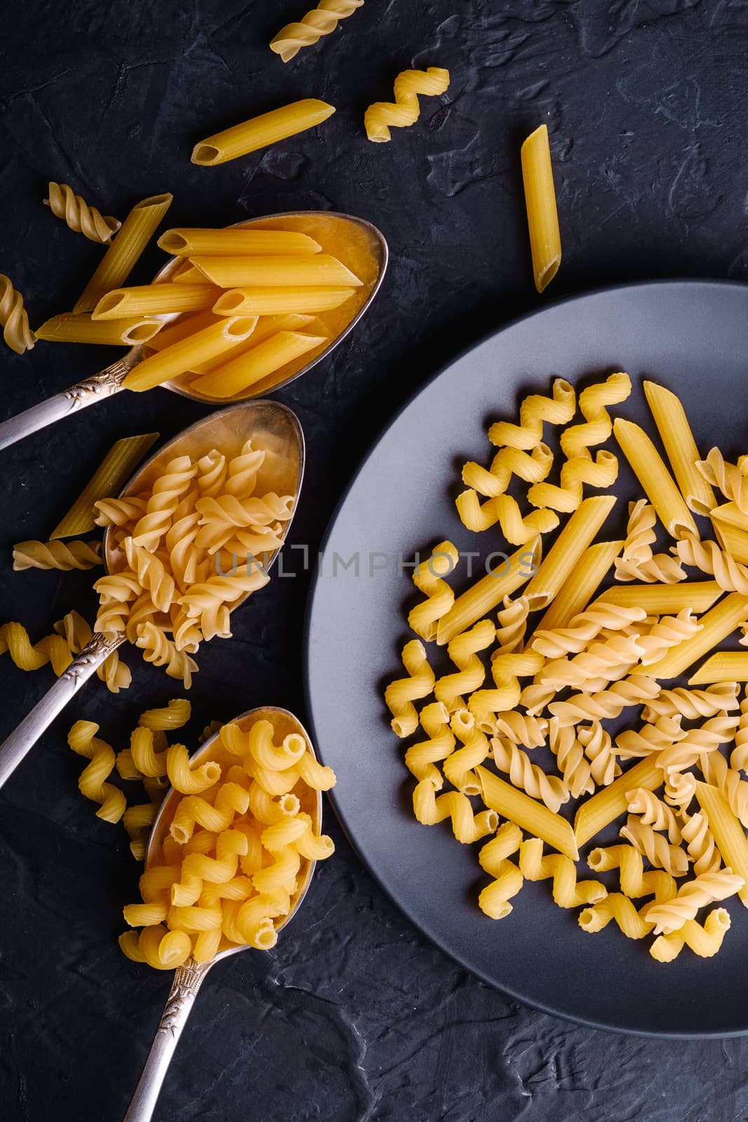 Three cutlery spoons and plate with variety of uncooked golden wheat pasta on dark black textured background, top view