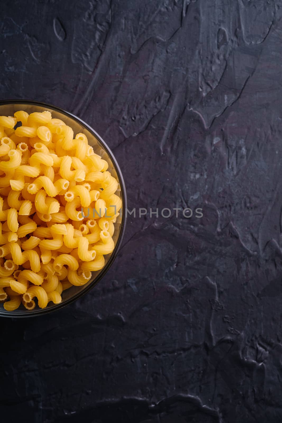 Glass bowl with cavatappi uncooked golden wheat curly pasta on textured dark black background, top view copy space