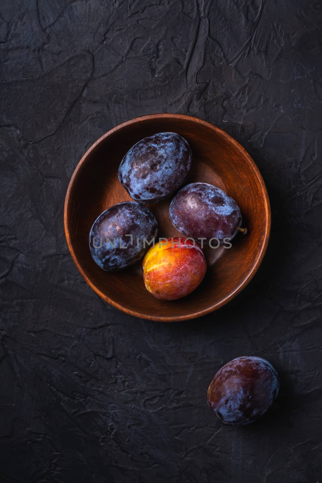 Fresh sweet plum fruits in brown wooden bowl, black textured background, top view