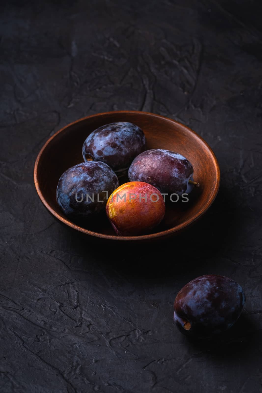 Fresh sweet plum fruits in brown wooden bowl, black textured background, angle view