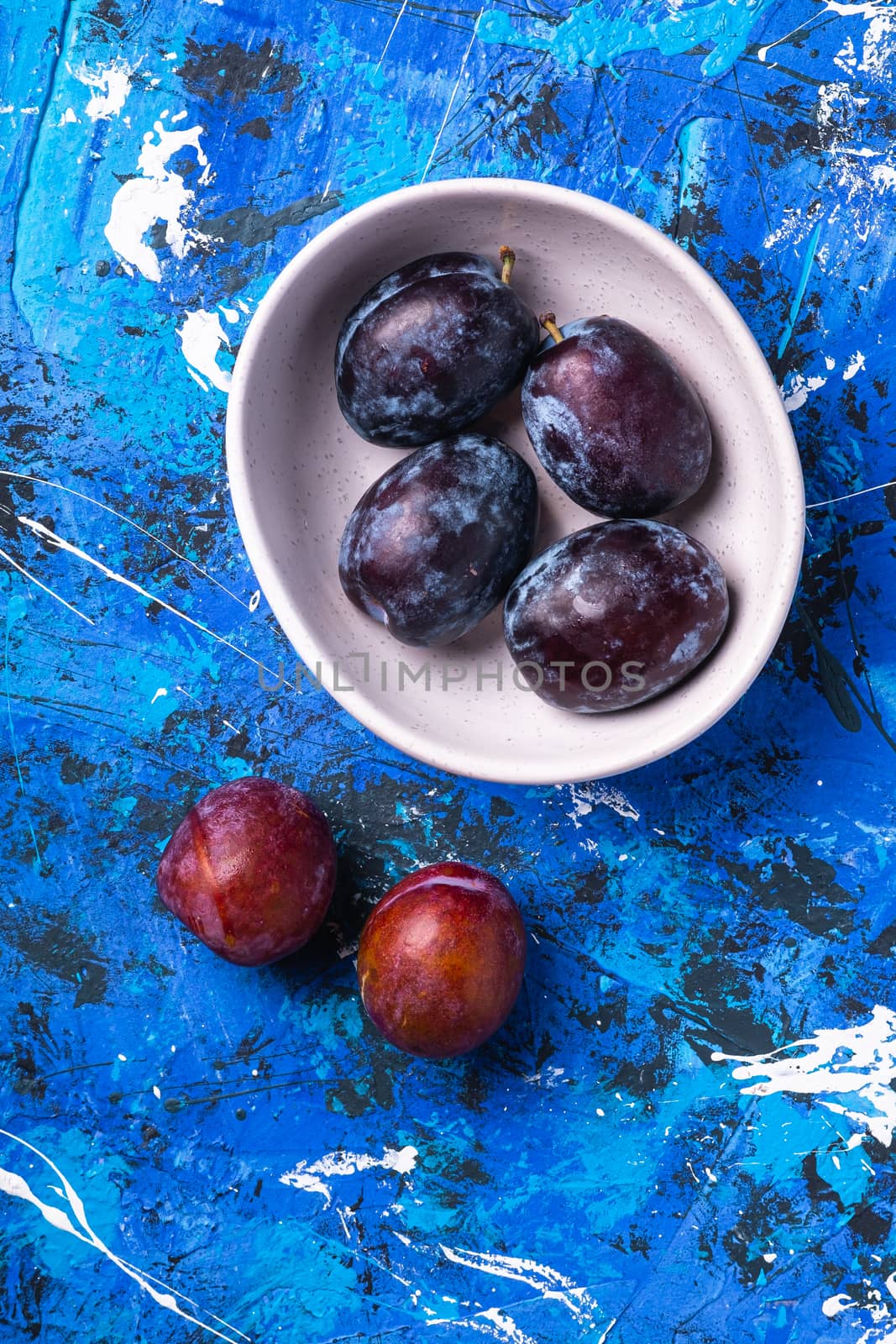 Fresh sweet plum fruits in white bowl on blue abstract background, top view