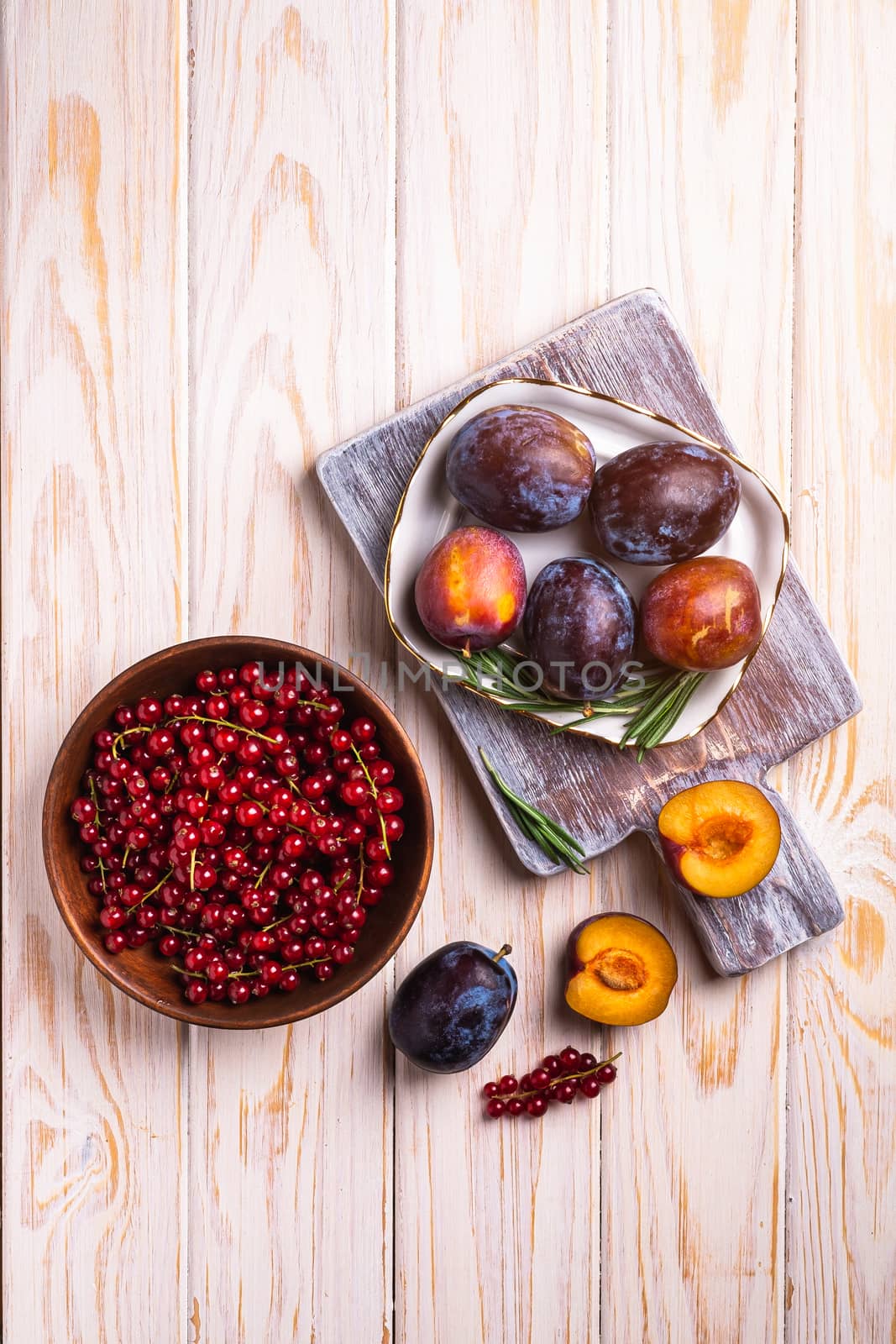 Fresh sweet plum fruits whole and sliced in plate with rosemary leaves on old cutting board with red currant berries in wooden bowl, wood table background, top view