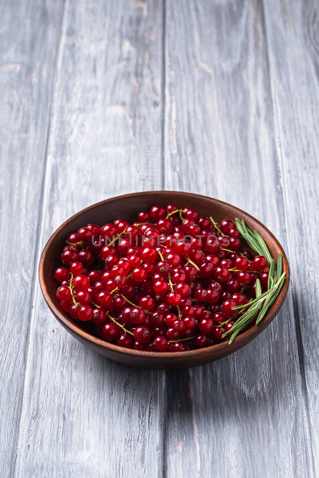Fresh sweet red currant berries with rosemary leaves in wooden bowl, grey wood background, angle view