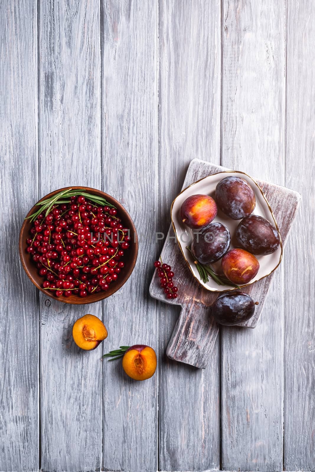 Fresh sweet plum fruits whole and sliced in plate with rosemary leaves on old cutting board with red currant berries in wooden bowl, grey wood background, top view copy space