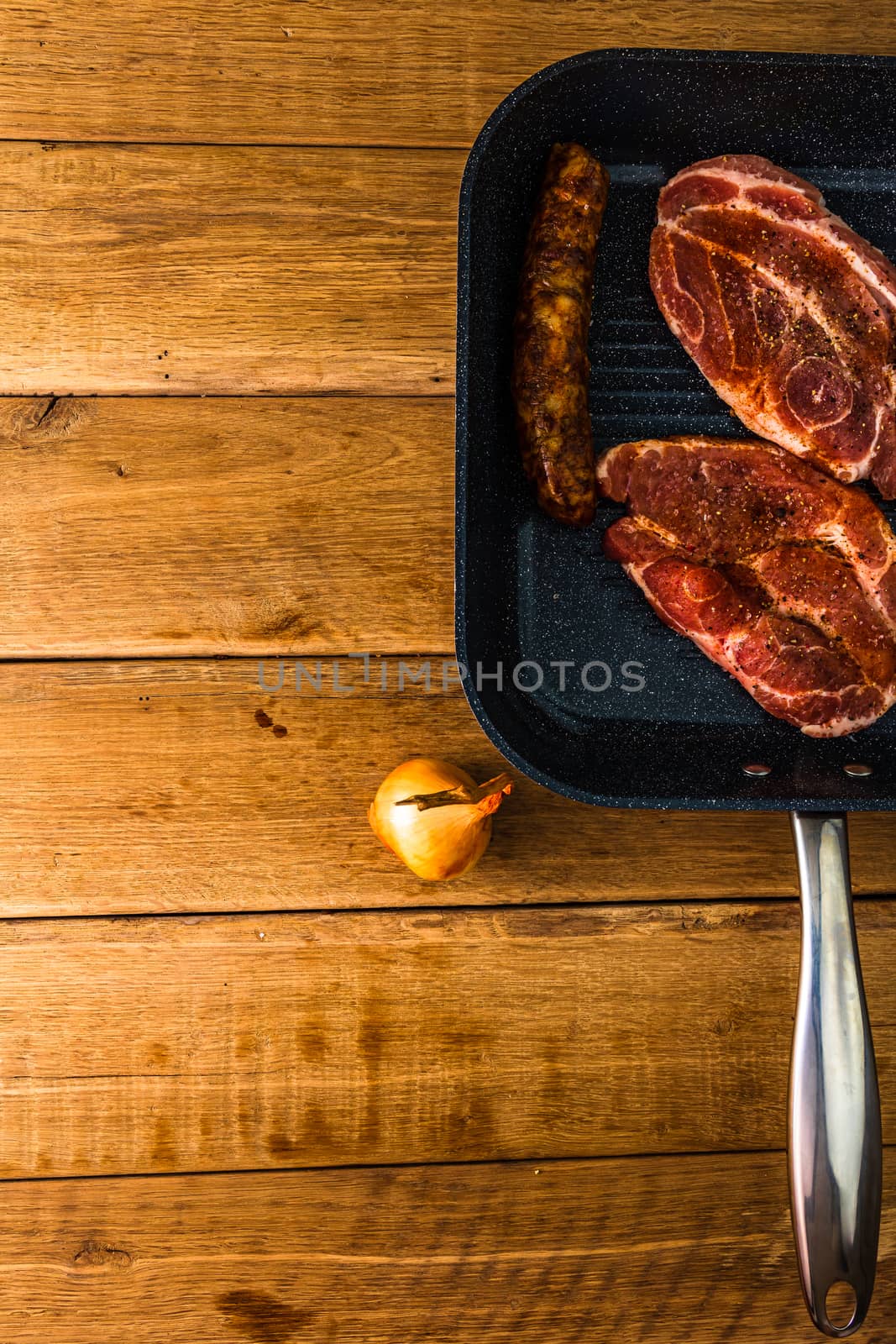 Pork chops with condiments and smoked sausage on a black frying pan, over wooden table, meat for bbq, top view, copy space, barbeque concept