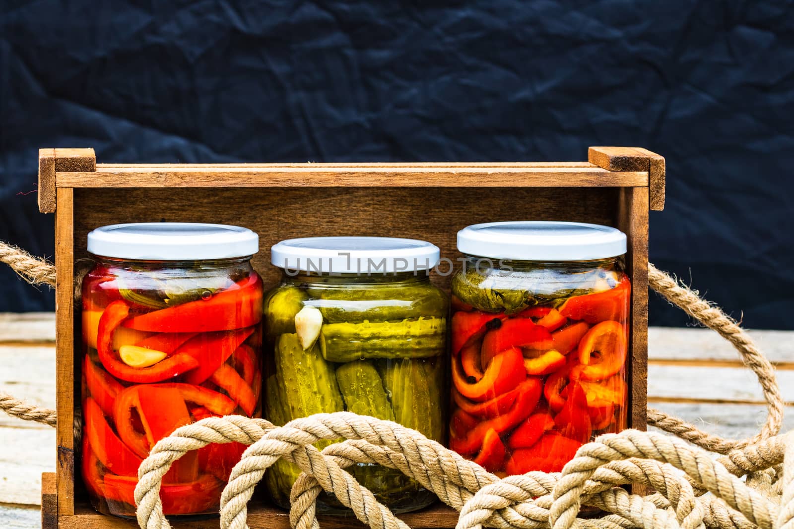 Wooden crate with glass jars with pickled red bell peppers and pickled cucumbers (pickles) isolated. Jars with variety of pickled vegetables. Preserved food concept in a rustic composition.