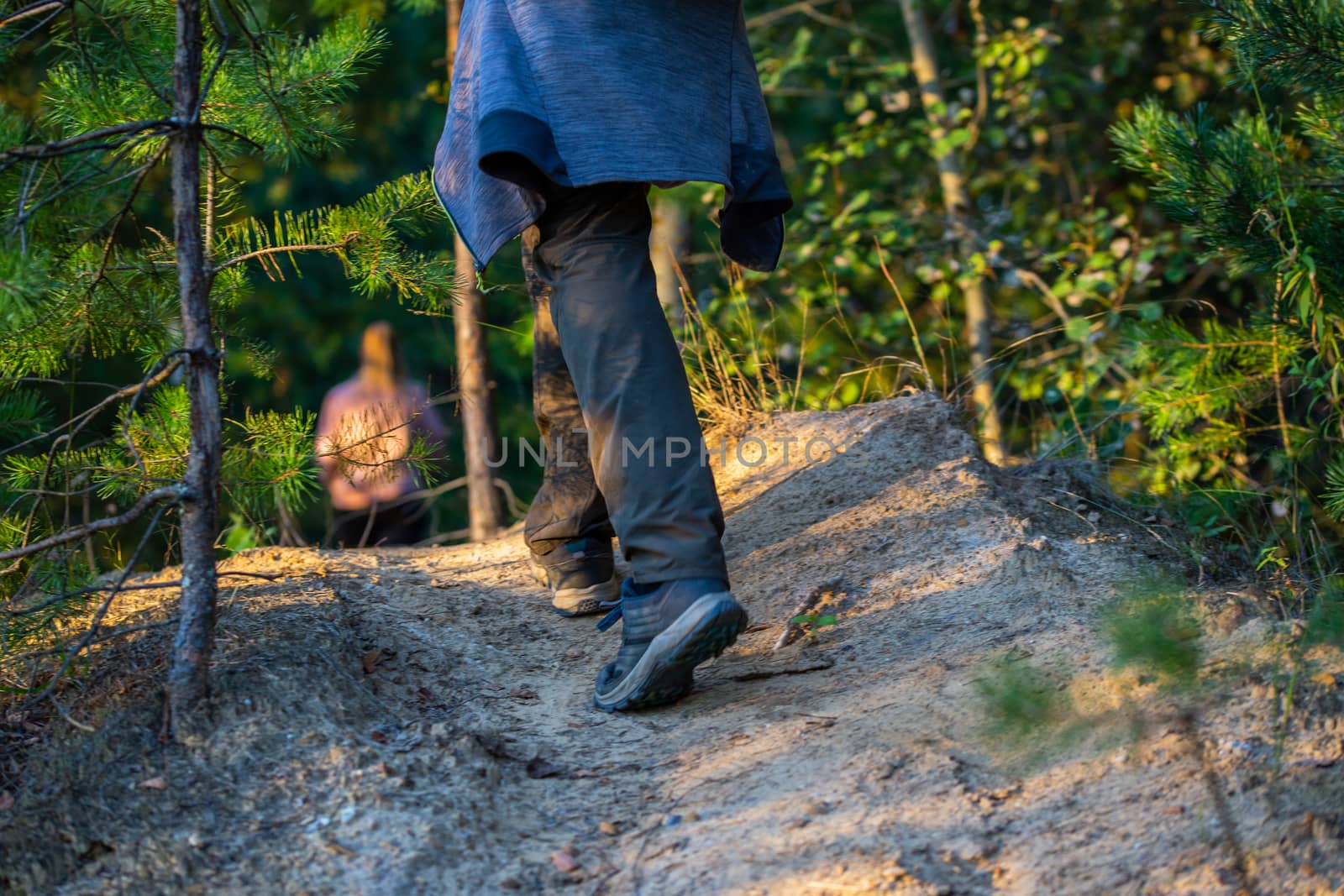 hiking man legs with shoes and gray pants somewhere in summer forest, close-up with selective focus. Blurred girl silhouette in the background.