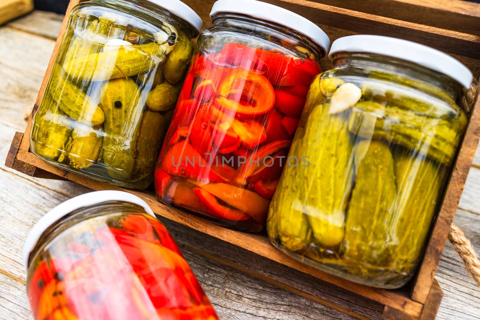 Glass jars with pickled red bell peppers and pickled cucumbers (pickles) isolated in wooden crate. Jars with variety of pickled vegetables. Preserved food concept in a rustic composition.