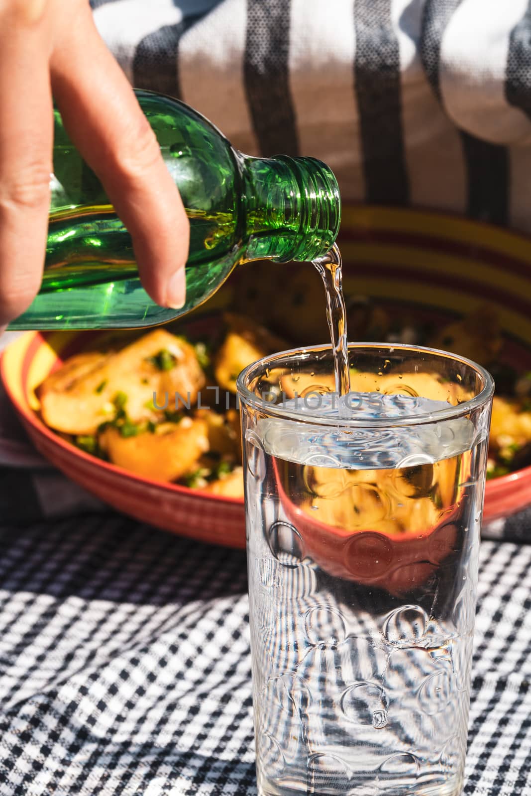 Pouring water from green bottle into a glass. Fried potatoes dish in the background