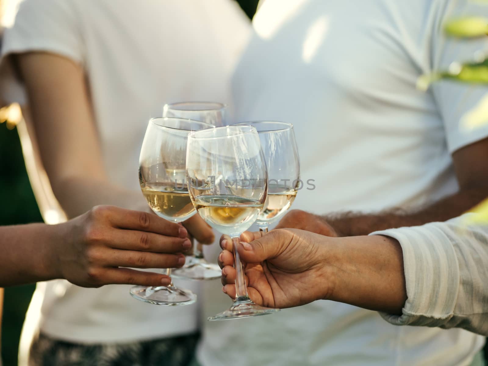 White wine glasses in hands. Four unrecognizable people clang glasses together outdoors.