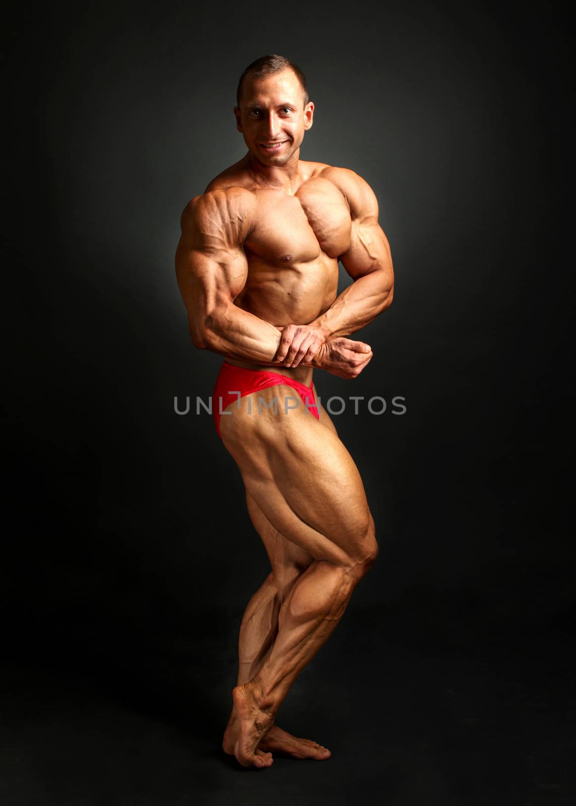 Studio shot of male bodybuilder posing, flexing and showing biceps and legs, with black background.