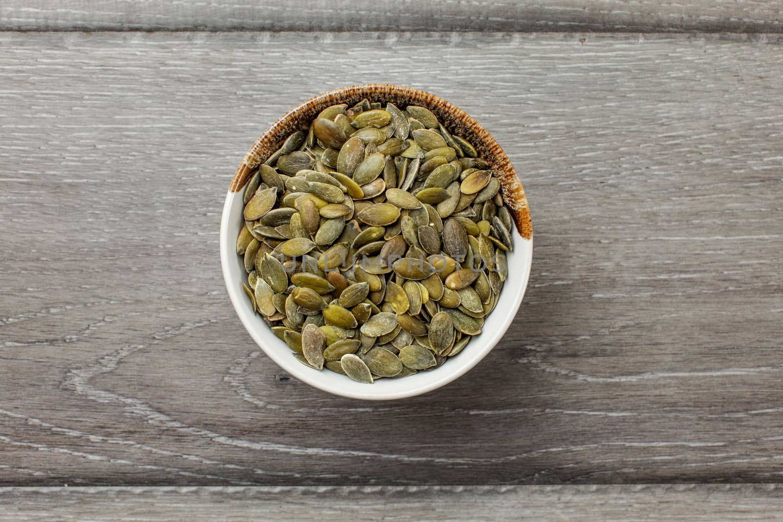 Table top view on small glass bowl full of roasted salted pumpkin seeds on a gray wood desk.