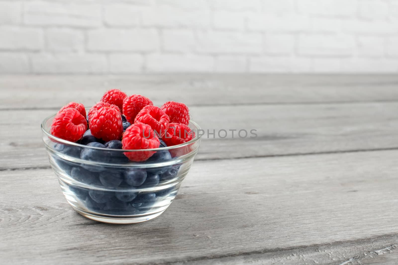 Small glass bowl full of blueberries and raspberries on gray wood table.