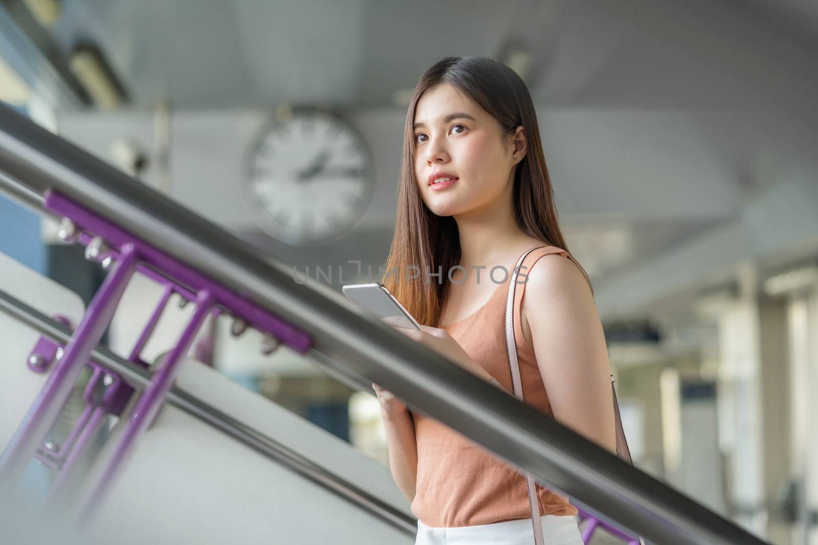 Young Asian woman passenger wearing headphone and using smart mobile phone and walking up the stairs in subway station when traveling in big city, commuter and transportation concept