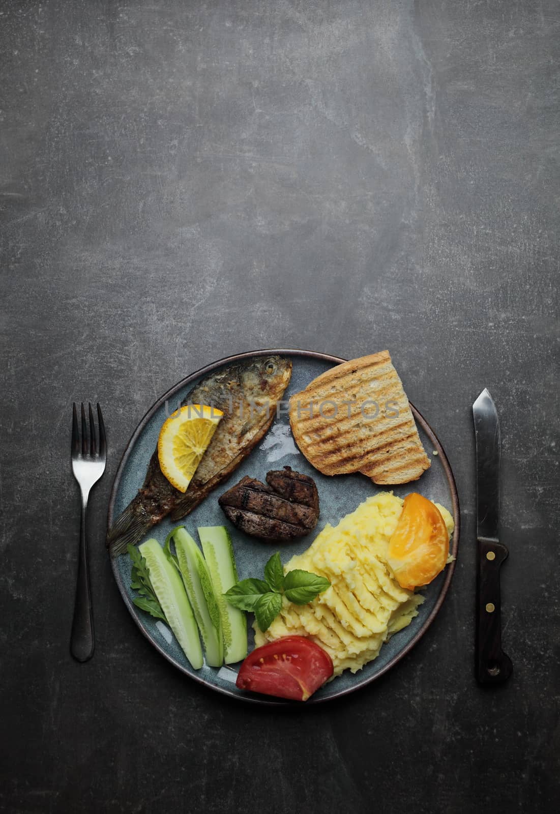 Fried fish, meat steak and vegetables on a plate. Concrete gray countertop by selinsmo