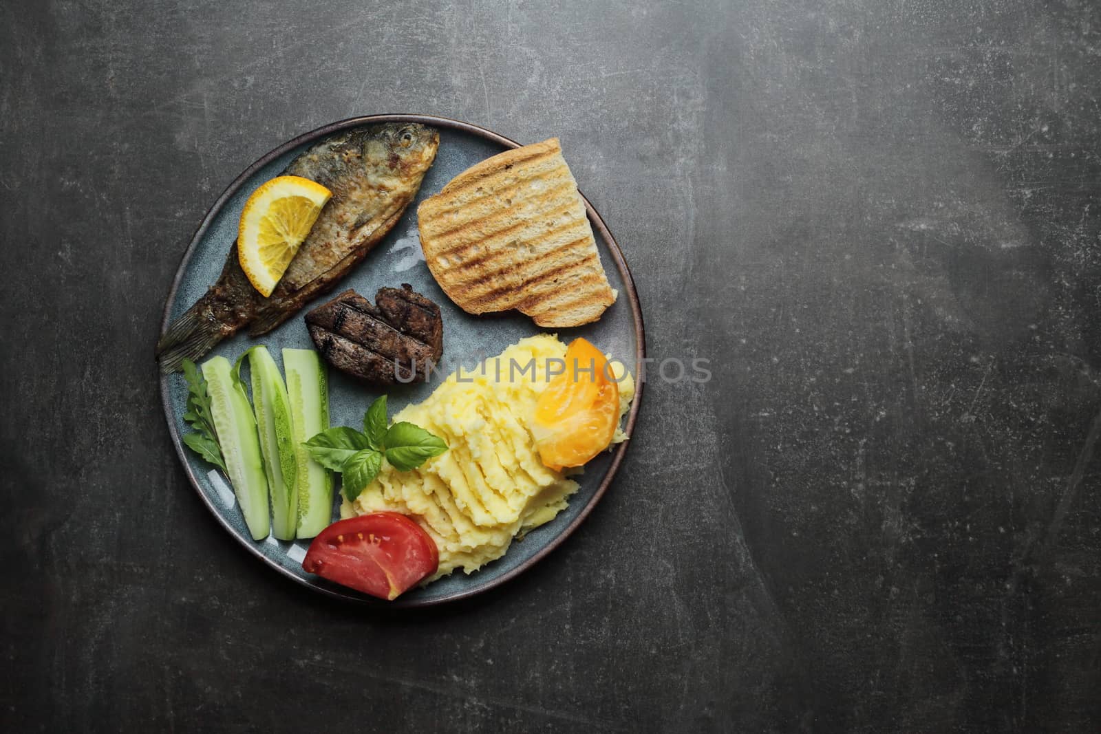 Fried fish, meat steak and vegetables on a plate. Concrete gray countertop by selinsmo