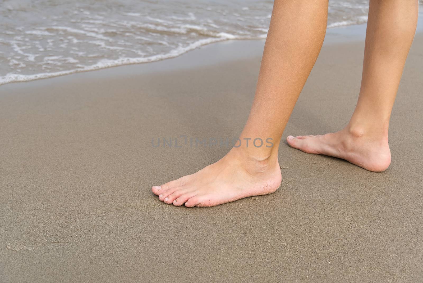 Close-up of child's legs walking on sand coast and sea water. Young girl legs on baltic sea beach. Teenager legs near sea line. by PhotoTime