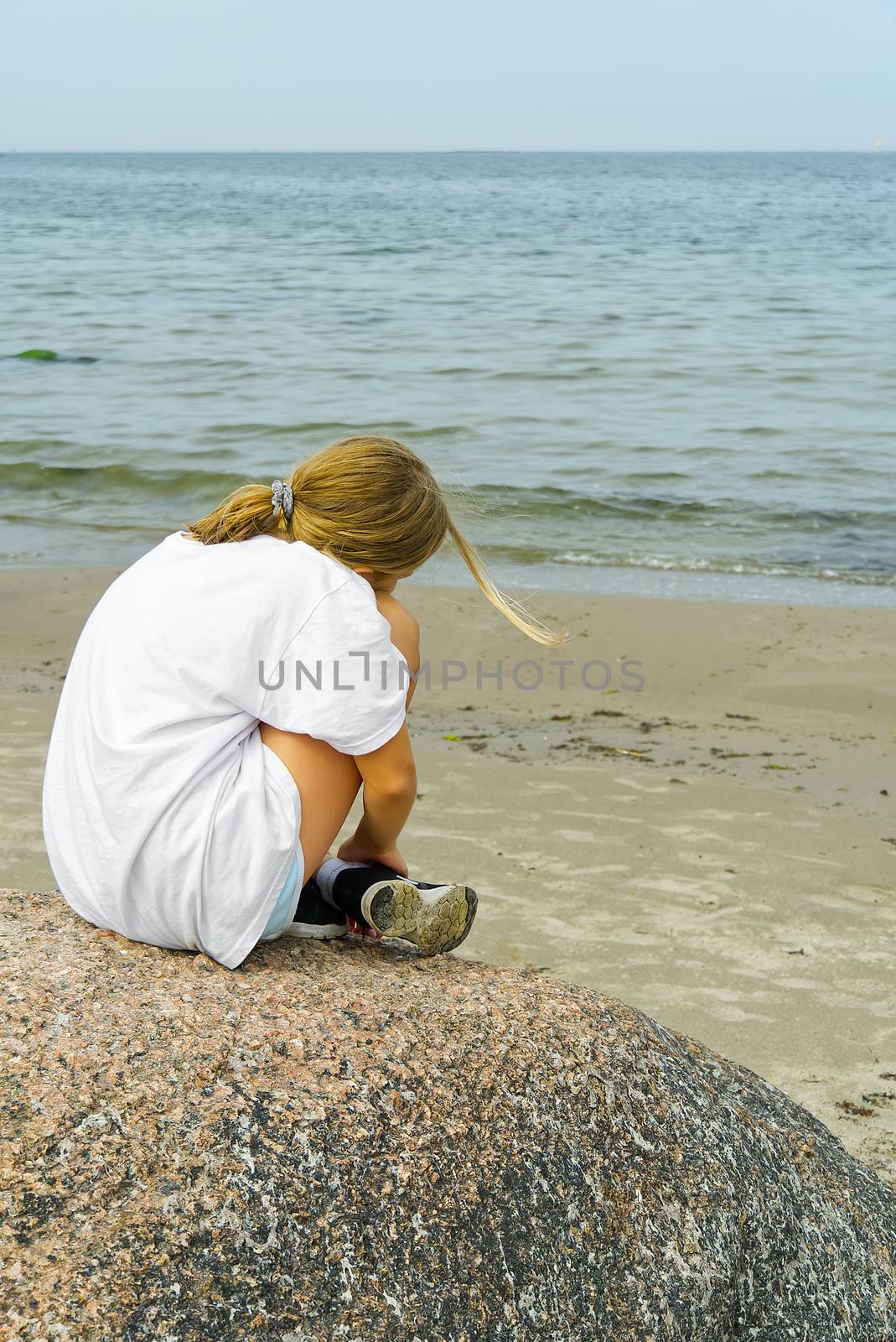 Side view of a charming little blond girl sitting on coastal stones and looking at the sea