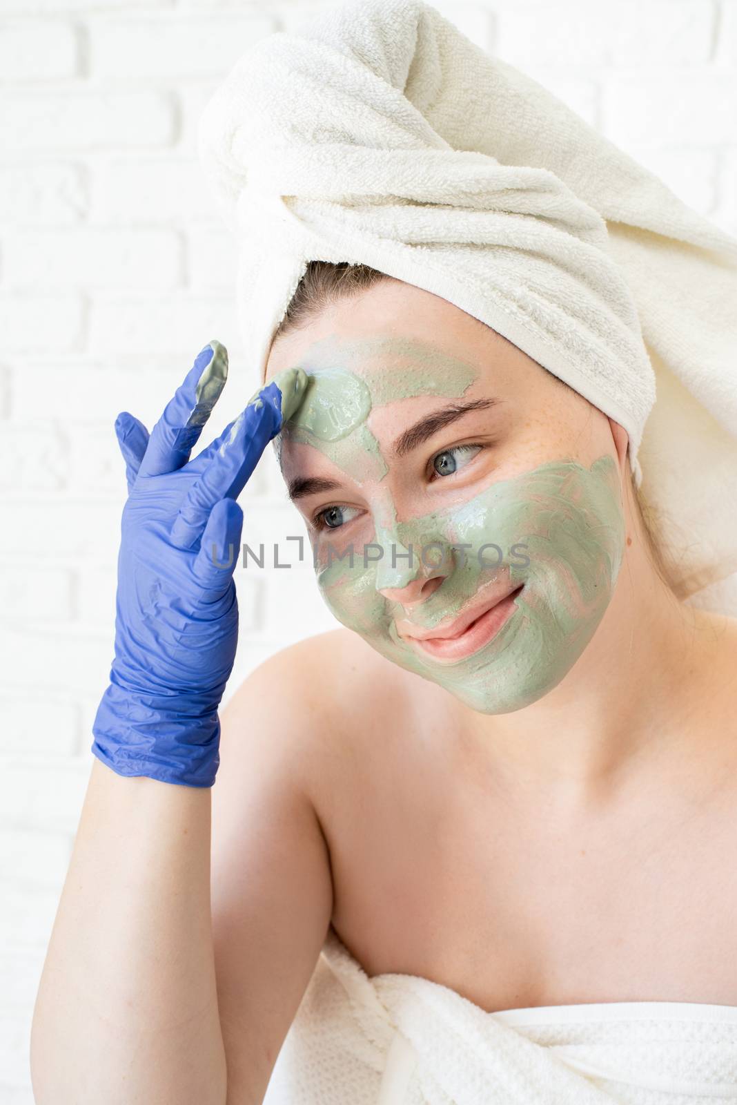 Spa and wellness. Natural cosmetics. Self care. Close up of happy young caucasian woman in white bath towels wearing gloves applying clay face mask looking at the mirror