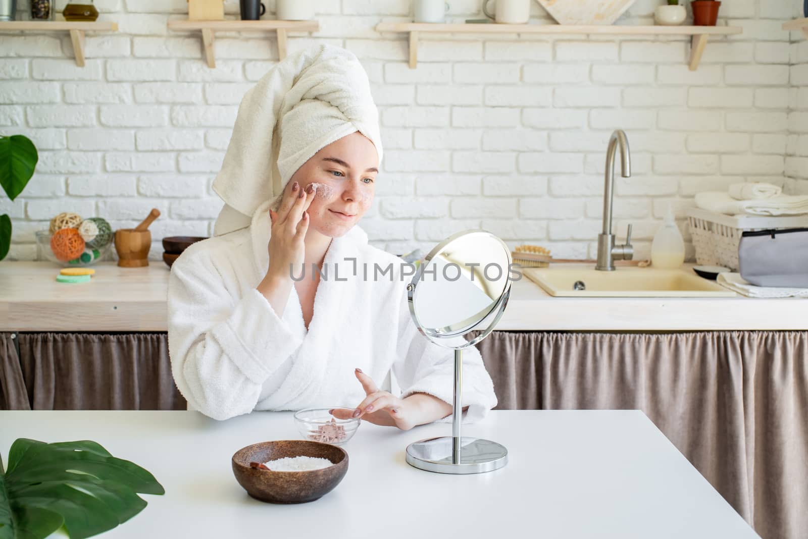 Spa and wellness. Natural cosmetics. Self care. Happy young woman applying face scrub on her face in her home kitchen looking at the mirror