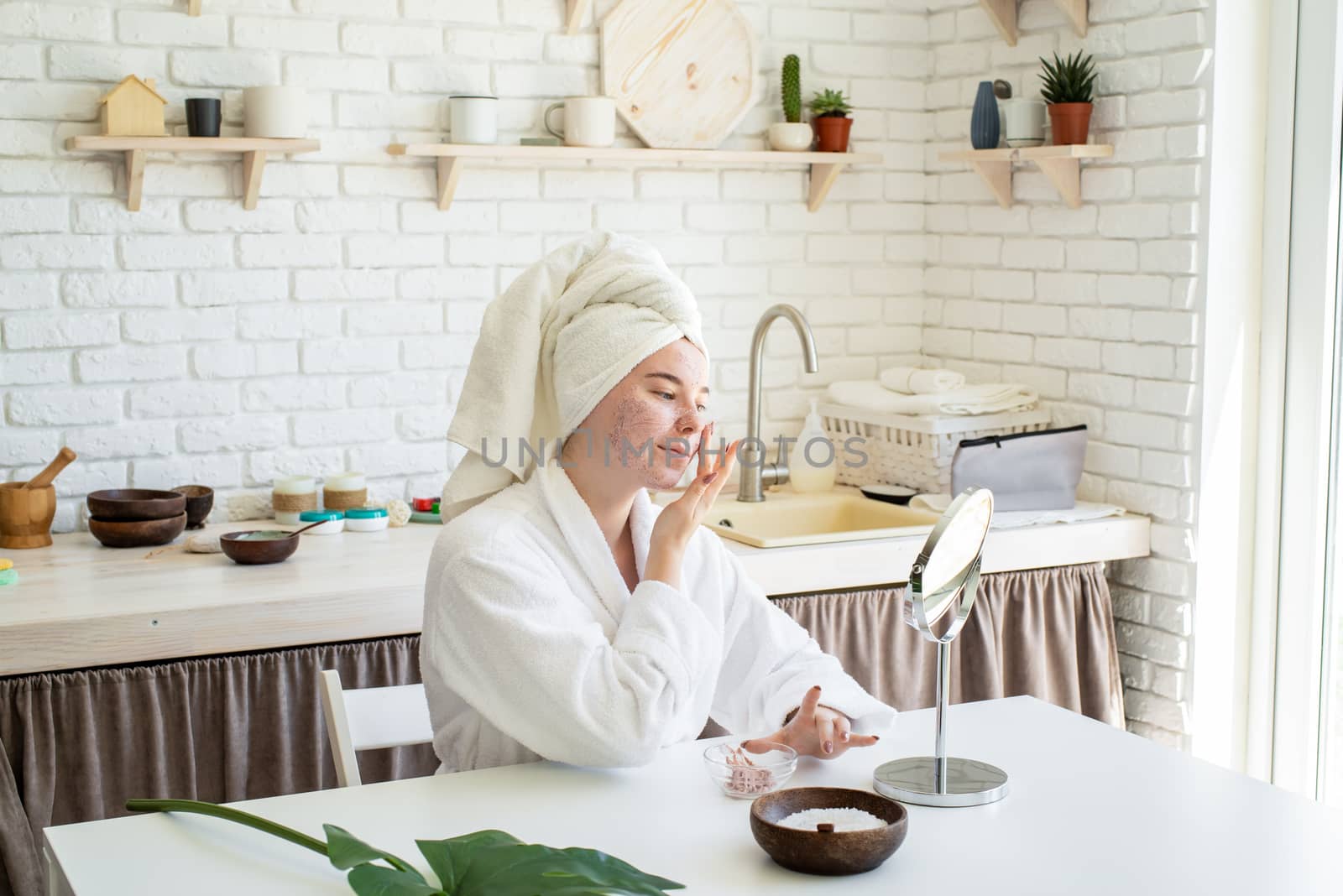 Spa and wellness. Natural cosmetics. Self care. Happy young woman applying face scrub on her face in her home kitchen looking at the mirror