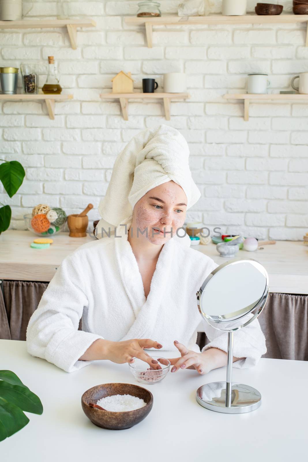 Spa and wellness. Natural cosmetics. Self care. Happy young woman applying face scrub on her face in her home kitchen looking at the mirror