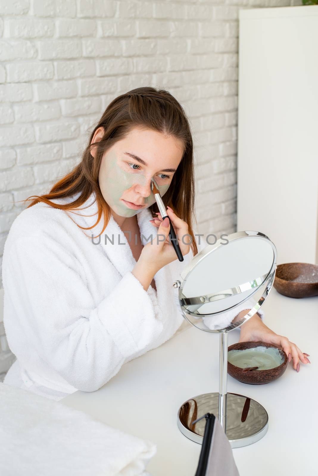 Spa and wellness. Natural cosmetics. Self care. Young smiling caucasian woman wearing bathrobes appplying clay face mask looking at the mirror