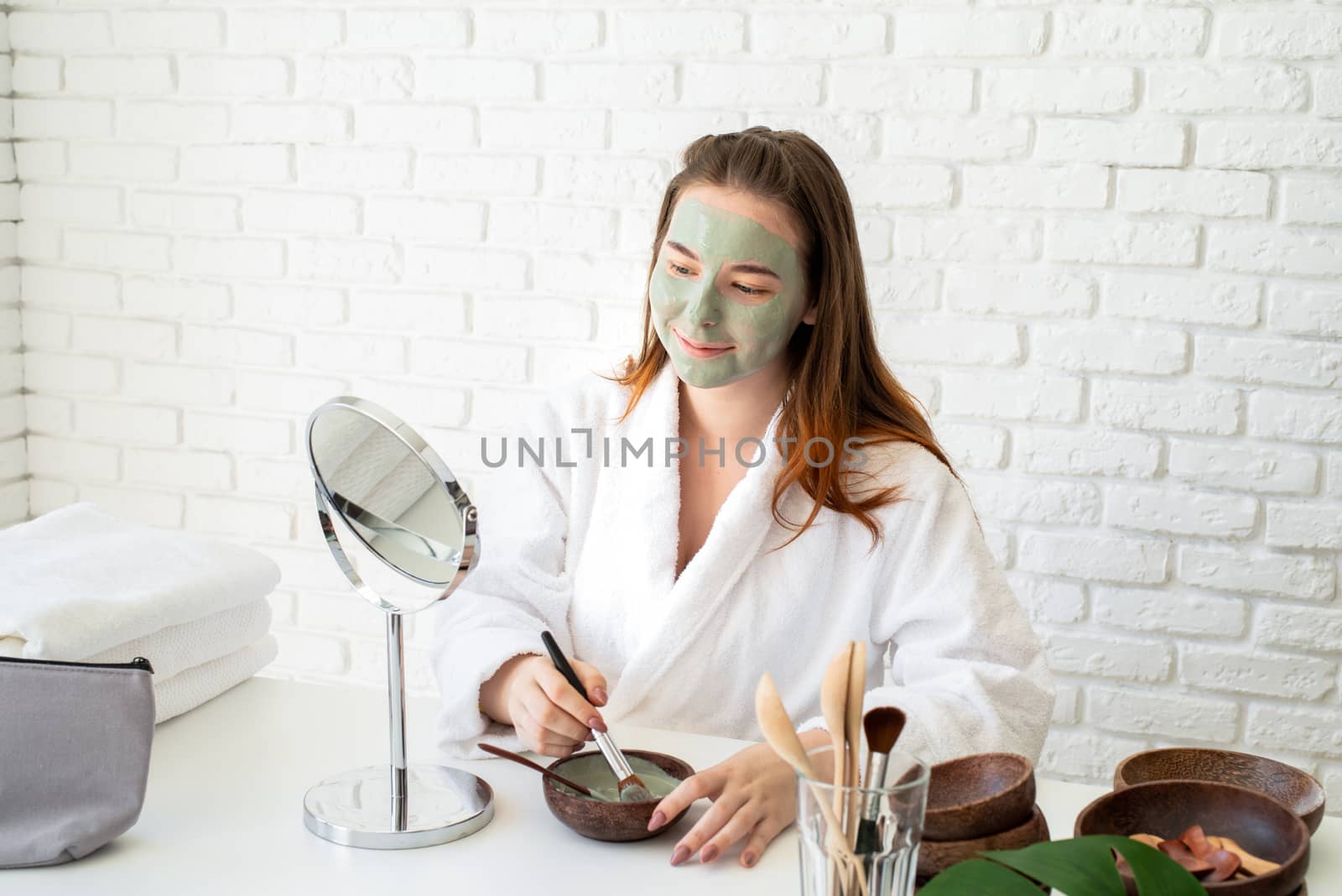 Spa and wellness. Natural cosmetics. Self care. Young smiling caucasian woman wearing bathrobes appplying clay face mask looking at the mirror