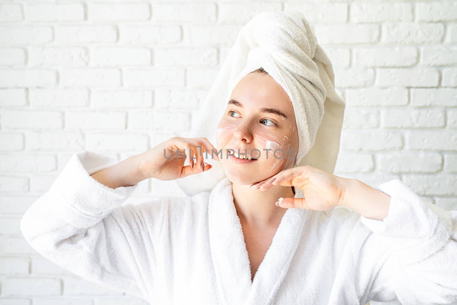 Spa and wellness. Natural cosmetics. Self care. Happy young caucasian woman in white bath towel applying face cream at home