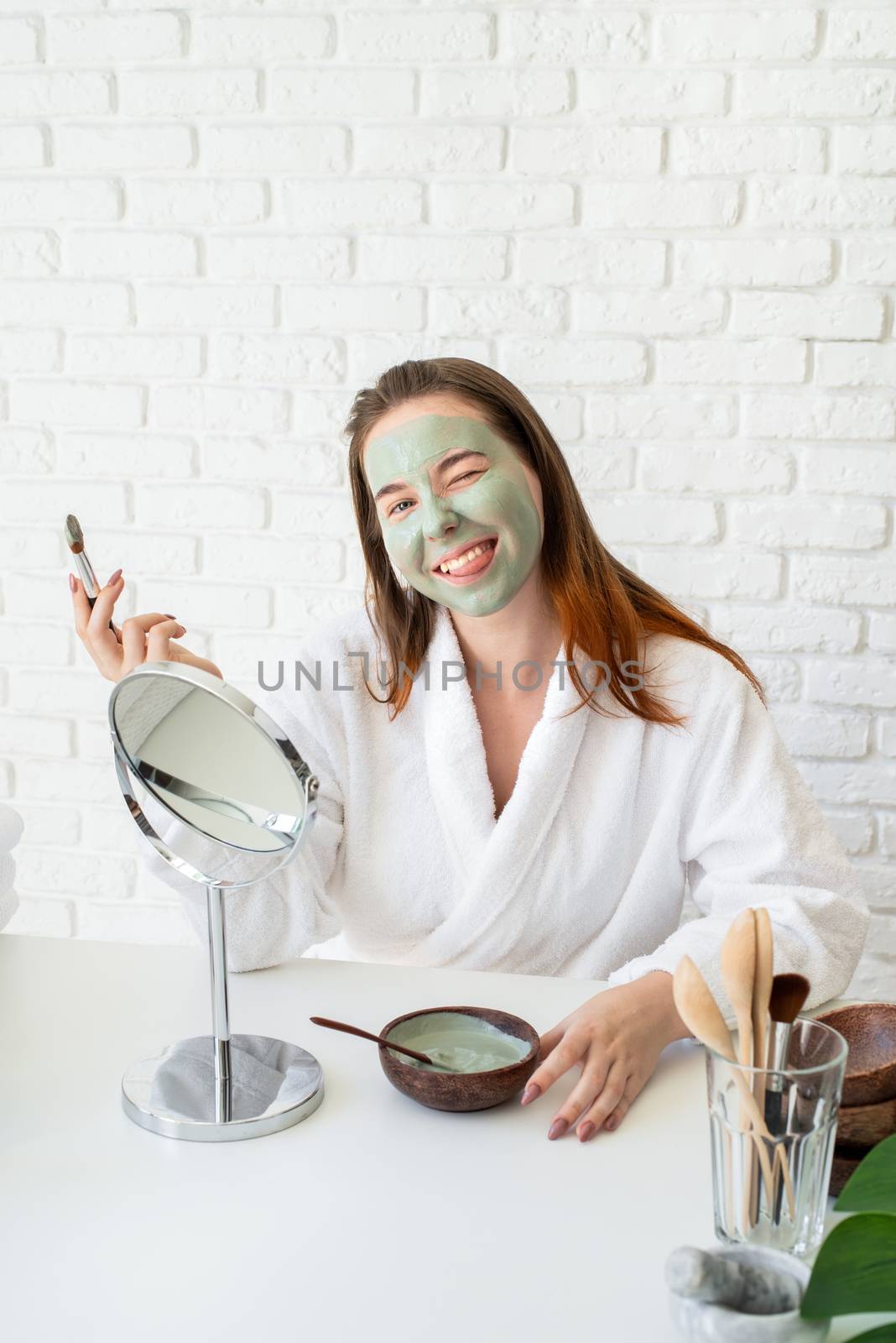 Young smiling caucasian woman wearing bathrobes appplying clay face mask looking at the mirror making funny face by Desperada