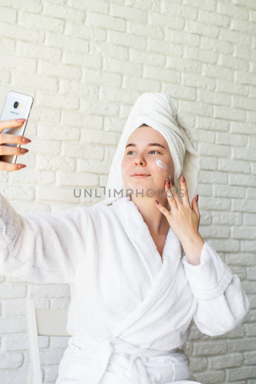 Spa and wellness. Natural cosmetics. Self care. Happy young caucasian woman in white bath towel applying face cream at home doing selfie