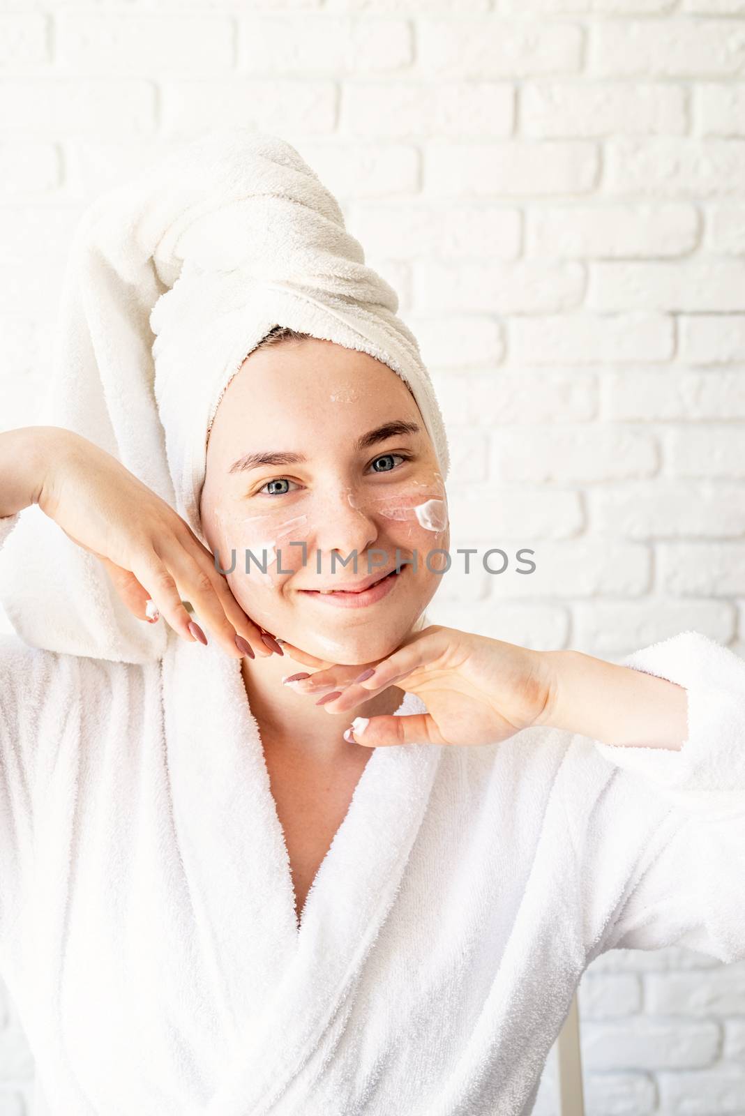 Happy young caucasian woman in white bath towel applying face cream at home by Desperada