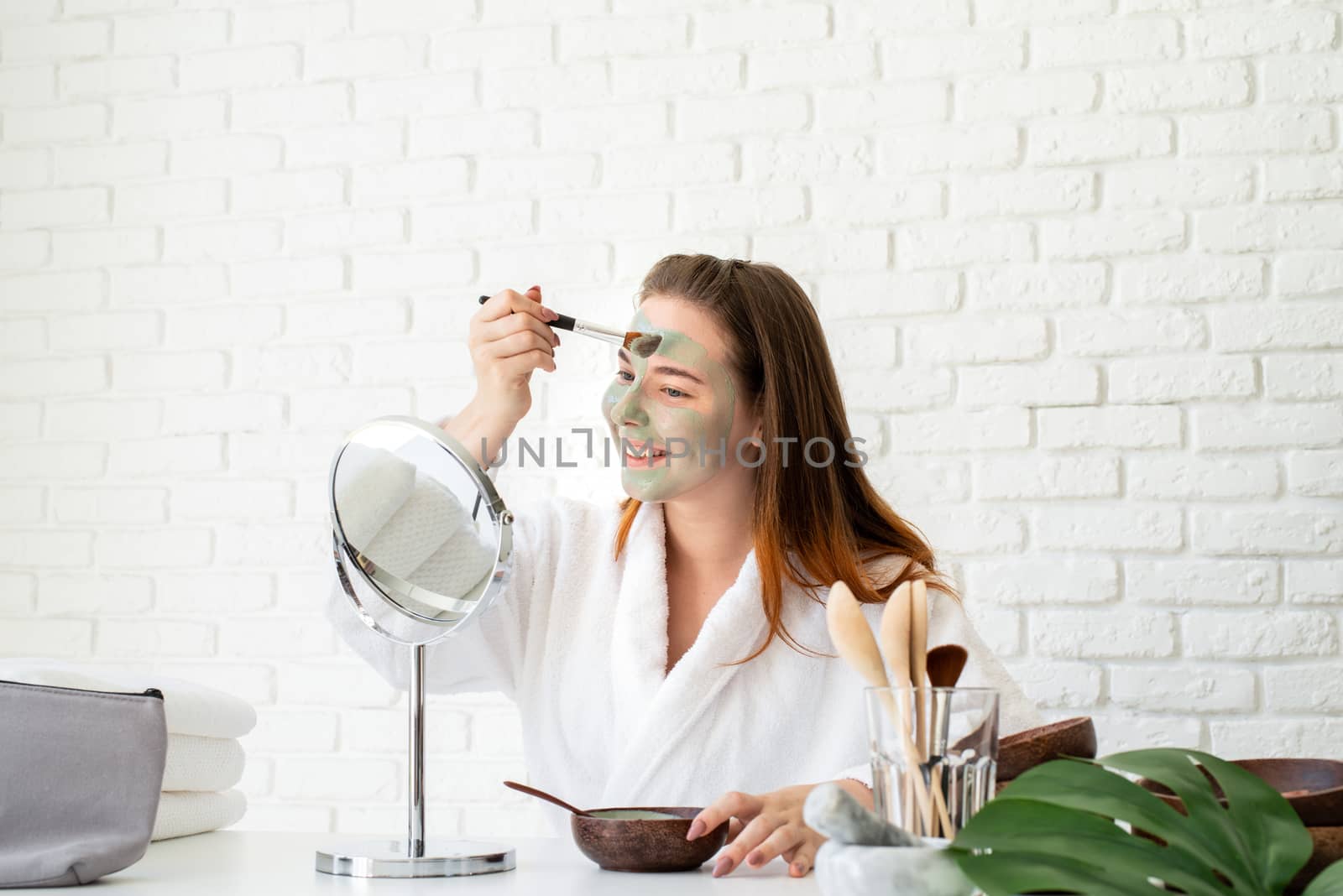 Spa and wellness. Natural cosmetics. Self care. Young smiling caucasian woman wearing bathrobes appplying clay face mask looking at the mirror