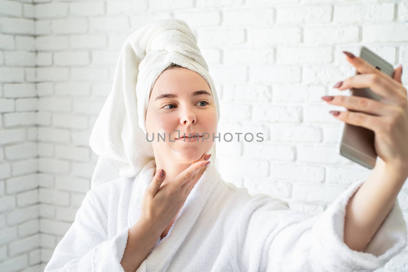 Spa and wellness. Natural cosmetics. Self care. Happy young caucasian woman in white bath towel applying face cream at home doing selfie