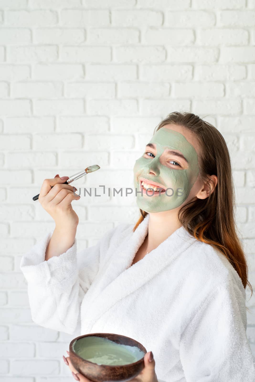 Spa and wellness. Natural cosmetics. Self care. Young smiling caucasian woman wearing bathrobes appplying clay face mask making funny face