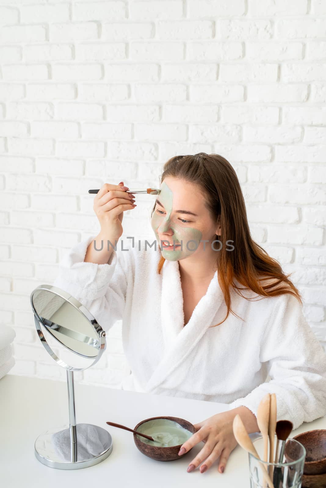 Young smiling caucasian woman wearing bathrobes appplying clay face mask looking at the mirror by Desperada