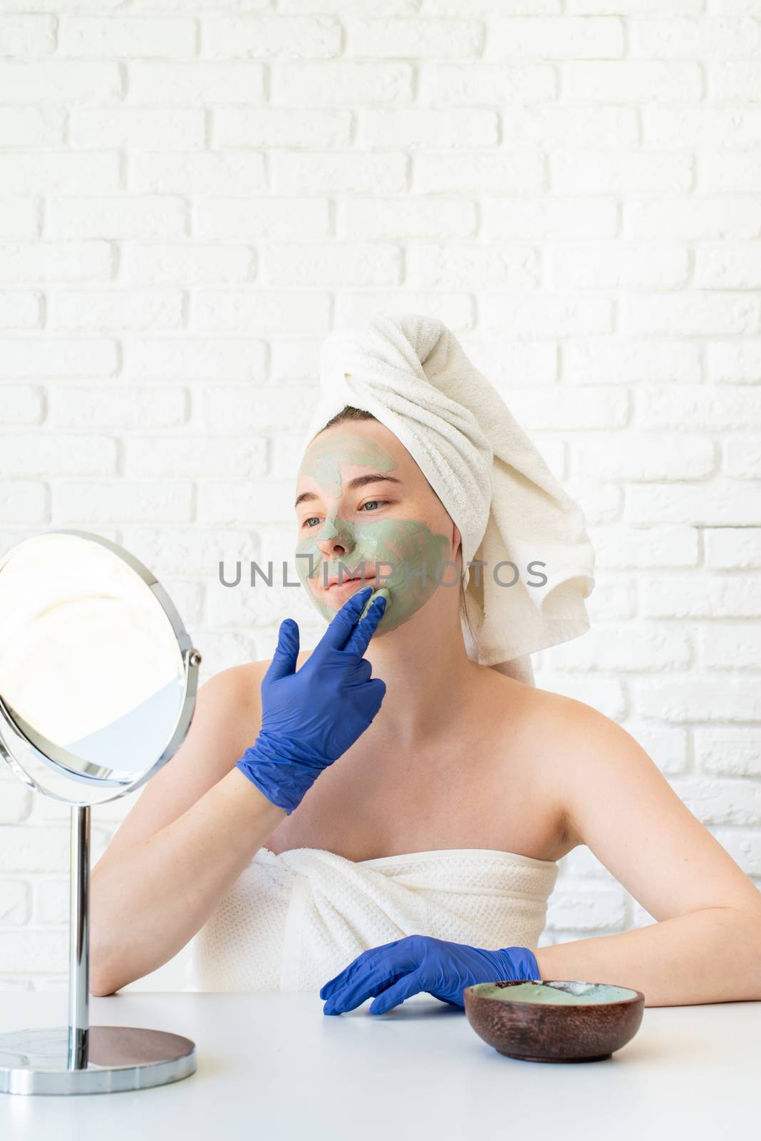 Happy young caucasian woman in white bath towels wearing gloves applying clay face mask looking at the mirror by Desperada